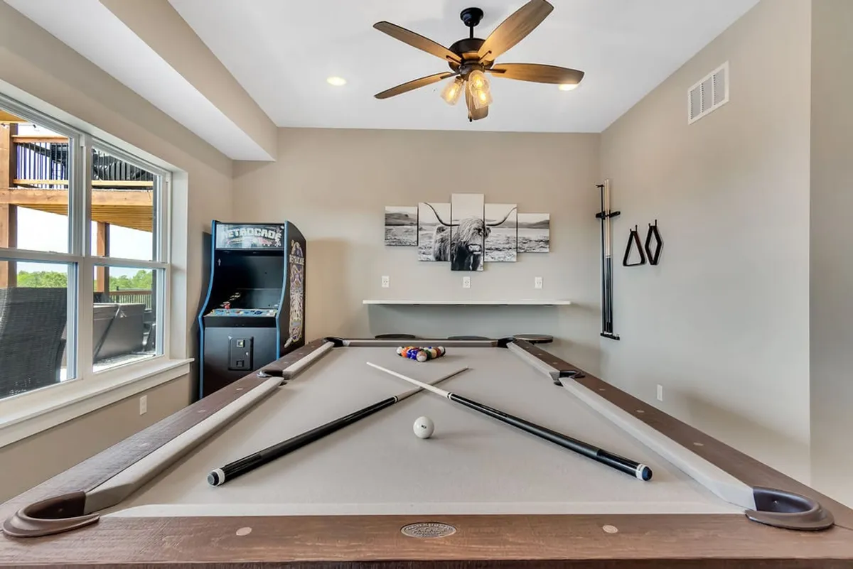A bright and inviting entertainment space with a pool table, Galaga arcade machine, wall art, and a ceiling fan. The room is arranged for socializing, playing games, and creating memorable experiences.