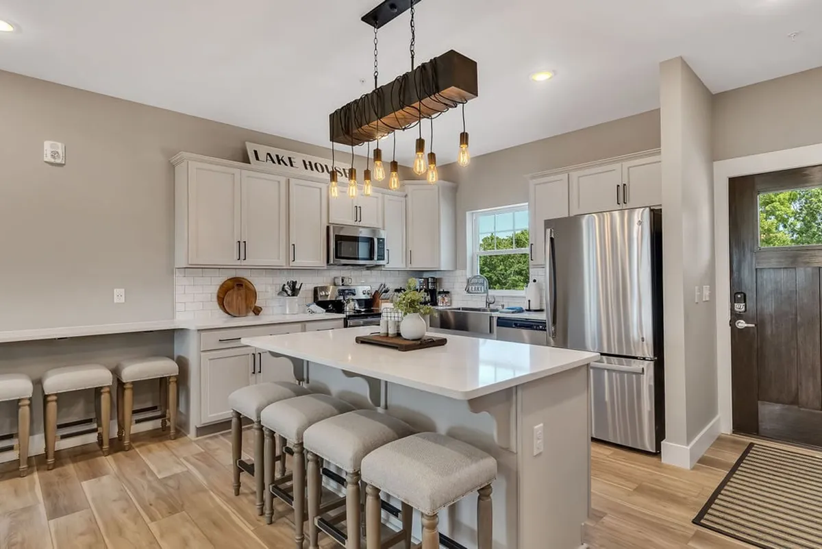 The image showcases a spacious, beautifully designed kitchen with a large island surrounded by comfortable seating, an elegant chandelier overhead, gleaming stainless steel appliances, and expansive windows offering tranquil lake views. The space is flooded with natural light, creating an inviting setting for family meals, entertaining, and gathering together.