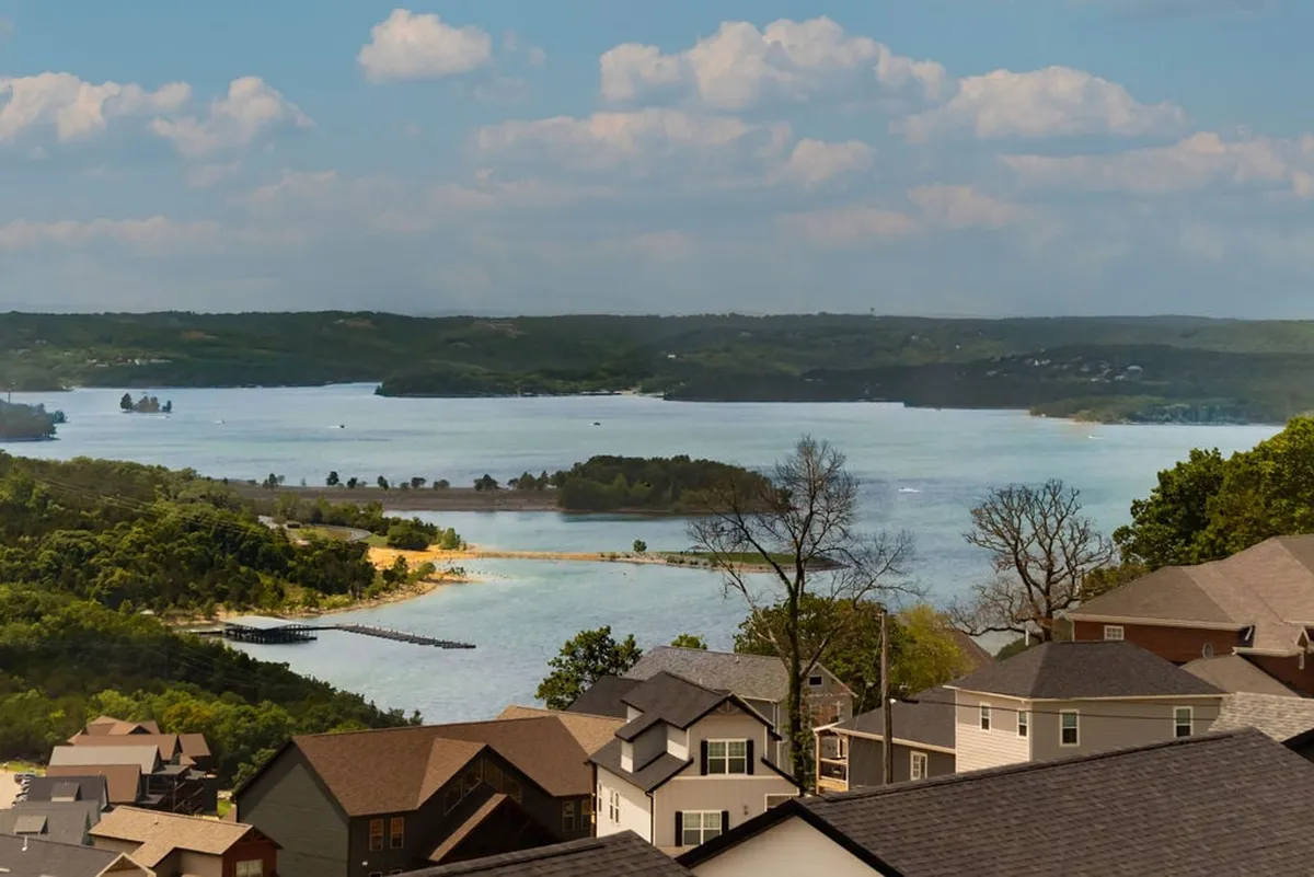 A sweeping view of the lake surrounded by rolling hills, dotted with houses and a lush tree line. The tranquil scene invites boating, fishing, hiking, and relaxing by the water, with the sun setting to cast a golden glow over the landscape.