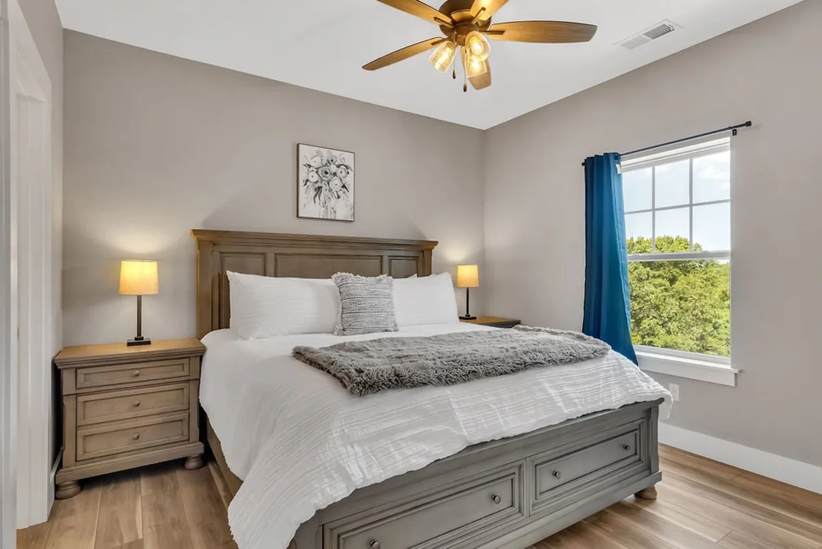 A tranquil king-sized bedroom featuring a plush bed with soft blue accents, matching nightstands with lamps, and a window with blue curtains that lets in natural light. Decorative artwork adds a calming touch to this inviting space, perfect for restful nights.
