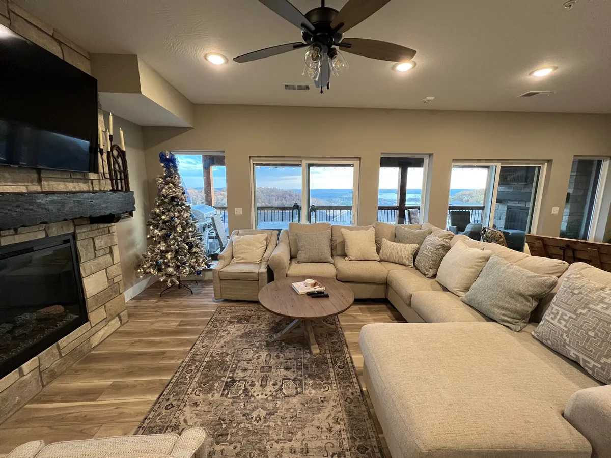 A spacious living room with a large sectional sofa, stone fireplace, and a beautifully decorated Christmas tree. Large windows offer scenic views, while the decor suggests a festive, welcoming environment for winter holidays.