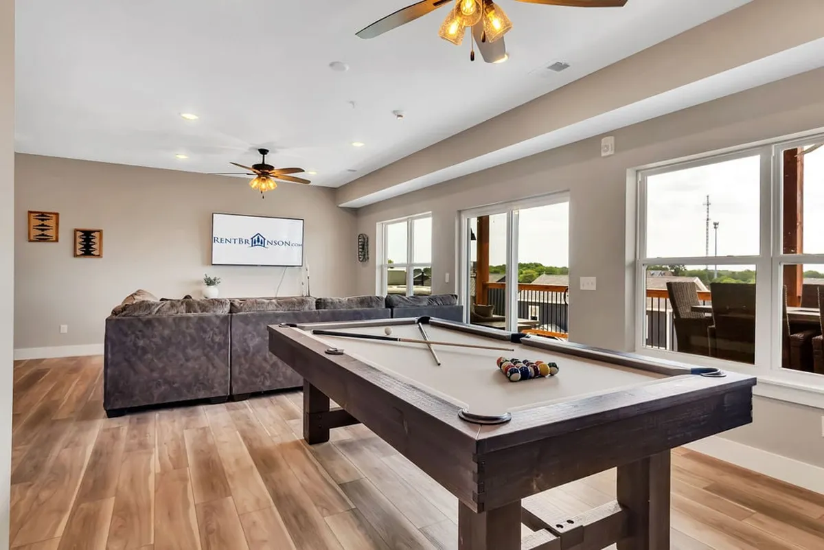 An interior view of a vibrant entertainment area complete with a pool table, sofa, large windows, and a TV. The space is designed for fun, offering options for playing pool, watching movies, and socializing, all in a relaxed and lively atmosphere.