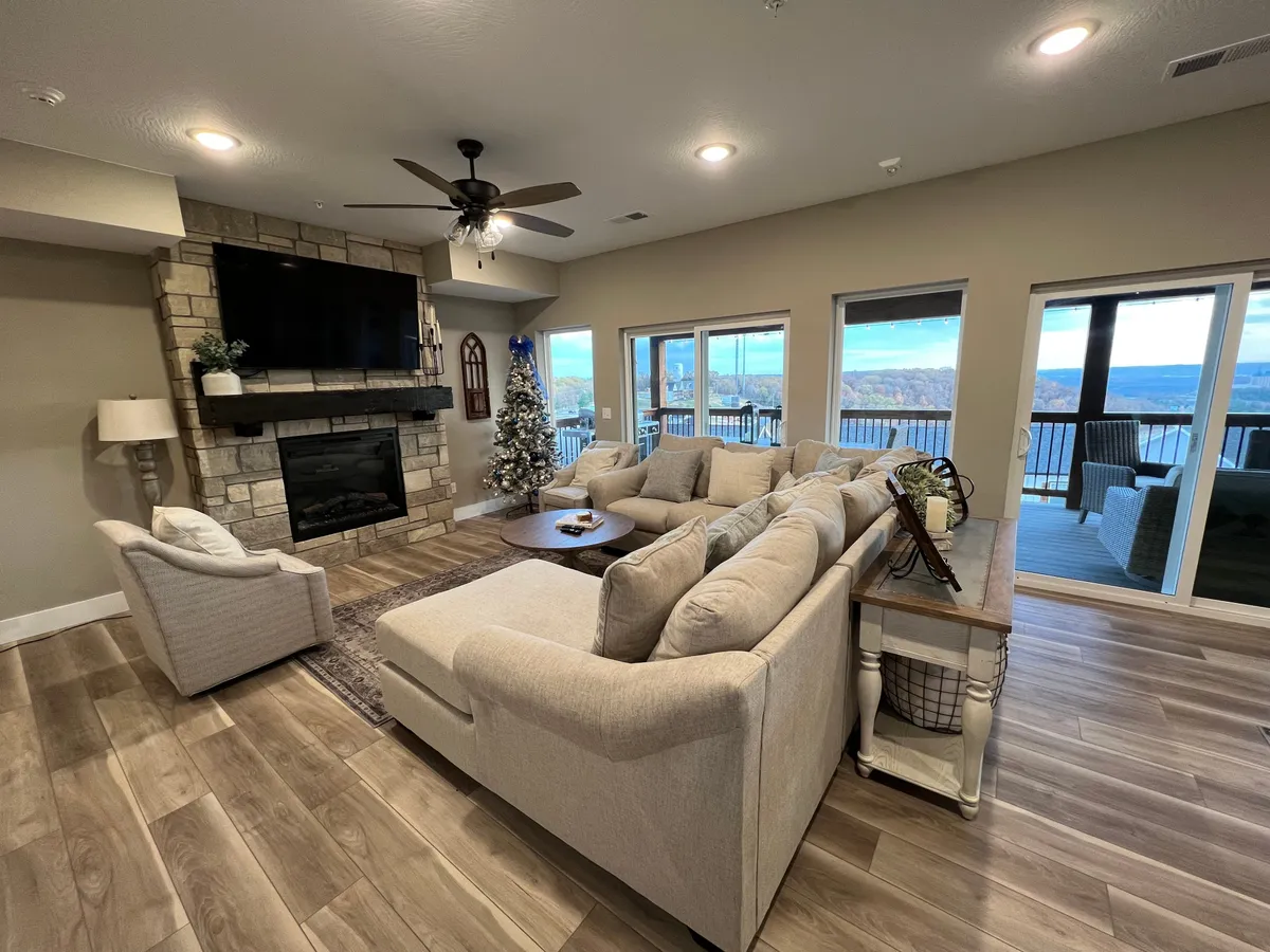 A spacious living area with a large sectional sofa arranged around a prominent stone fireplace. Expansive windows provide scenic views and natural light, while the warm, rustic furnishings create an inviting atmosphere for group relaxation and conversation.