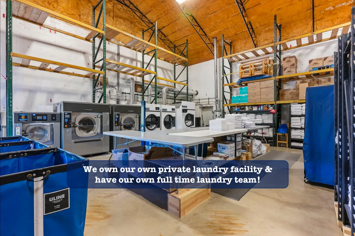 A utilitarian interior space with industrial laundry machines, organized shelves of supplies, and a folding table. The mood is efficient and professional, emphasizing cleanliness and care in property management.